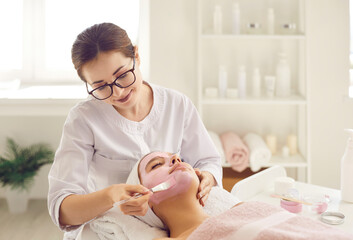 Beautician at beauty parlor applying soothing pampering facial mask with pink kaolin clay on young...