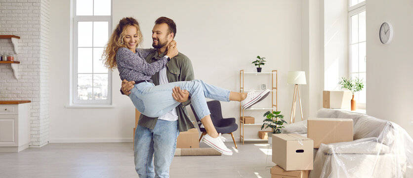 Joyful couple moves to their own first home. Happy loving husband holding his beloved wife in his arms in their new house during move. Property buying or mortgage concept. Banner.
