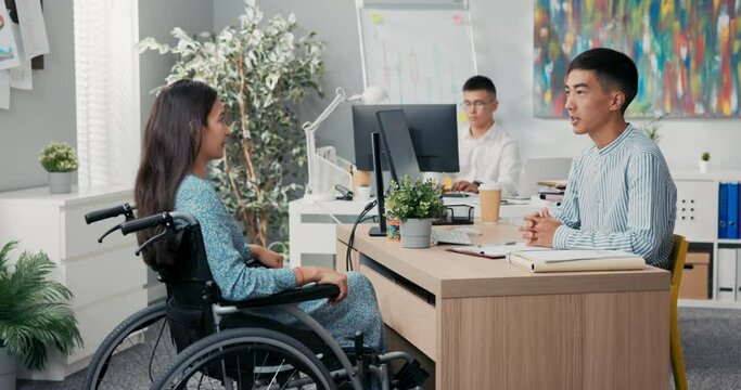 Woman is sitting in wheelchair at desk in office, across from her employer is holds out hand, they are shaking hands thanking each other for interview congratulating greeting saying goodbye