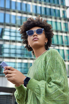 Outdoor Shot Of Curly Haired Woman Wears Sunglasses And Green Jumper Uses Smartphone While Walking At Street Among Modern Glass City Building. Female Blogger Spends Free Day Outside Uses Internet