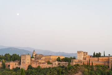 Obraz premium Landscape view of the famous Alhambra at sunset, Granada, Andalusia, Spain. White moon on the left. Pale blue sky on the background. With copy-space.