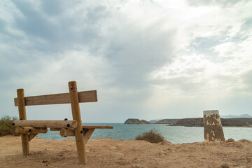 Wooden bench at the top of a hill, on a viewpoint, with blue mediterranean waters. The famous "Cuatro Calas" (meaning: "Four Coves") are visible in the distance. Aguilas, Murcia, Spain.