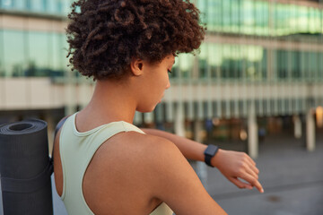 Sideways shot of sportive curly haired woman trainer checks time on wearable smartatch walks outdoors in urban setting returns from yoga training with karemat wears casual comfortable clothes