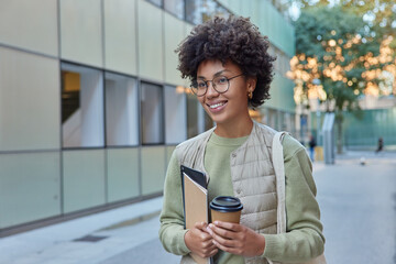 Fototapeta na wymiar Happy female student with curly bushy hair walks on street drinks takeaway coffee holds notepad and modern tablet smiles joyfully and looks forward poses in urban setting goes to university.