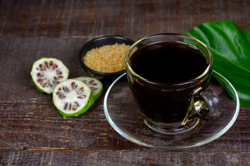 Noni juice in transparent glass cup and fresh noni fruit with green leaf on rustic wooden background.
