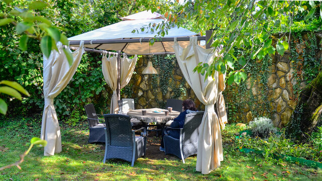 .In the garden, among the trees, there is an awning gazebo against a stone wall. The sun, breaking through the foliage, lies on the table, the grass, shines through the roof. 
