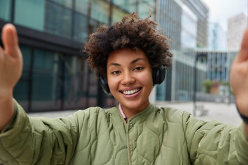 Positive curly haired woman smiles happily makes selfie listens music via headphones wears jacket poses outdoors against urban background spends free time in big city. People lifestyle hobby
