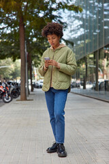 Vertical shot of female travel blogger checks route on location application reads received message during mobile mailing wears jacket jeans and shoes stands on pavement in city makes online booking