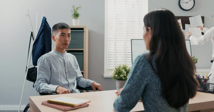 Disabled man dressed in smart blue striped shirt sits at desk in corporate office in wheelchair, he is applying for secretarial job, female corporate executive is conducting interview she ask question