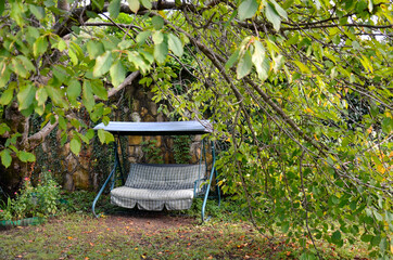 In the garden, against a stone wall, there is a sofa swing. The branches of the trees fell like a tent over the swing. The sun illuminates the leaves.