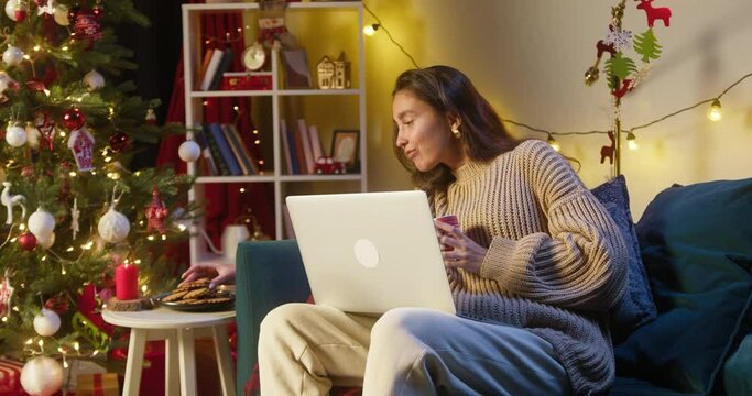 Young woman watching film on laptop, sitting on sofa in living room. Female student watches movie on computer, eating cookies and drinking. New year time, christmas holidays and lockdown.