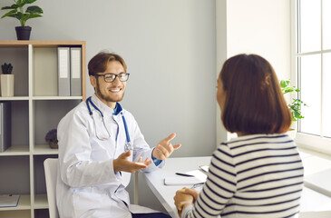 Smiling friendly male doctor tells his patient good news at meeting in hospital office. Unrecognizable female patient sitting with her back to camera talking to her family doctor. Healthcare concept.