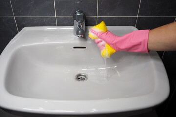 A woman's hand in a rubber pink glove, a yellow sponge with soapy foam, a white washbasin. Household and cleanliness concept
