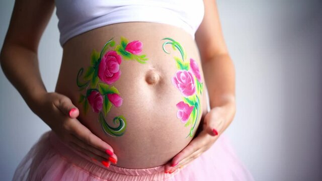 A close-up of the abdomen of a pregnant woman on a light background, the girl strokes the pregnant stomach. On a pregnant belly painted aqua makeup pink flowers with green leaves. High quality FullHD