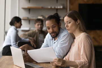 Concentrated caucasian female employee holding paper document, discussing research results with skilled male african american colleague, working on project presentation or data statistics on computer.