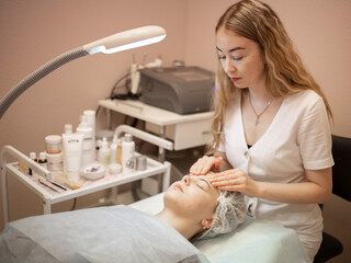 A girl-cosmetologist in a white coat, in a medical office, performs a procedure for a client,...