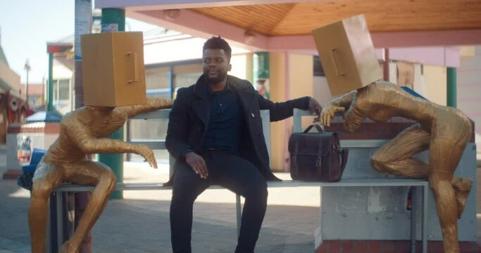 Handsome African business man sits between abstract statues on a bench in a city as he looks at them and then starts laughing.