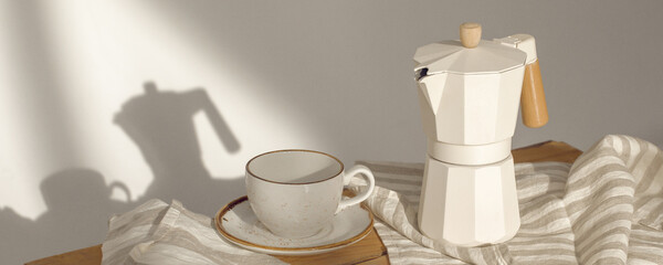 Italian coffee maker with wooden handle and white coffee craft cup on solid wood stamp decorated with tea towels in creamy tones on sunlight creating beautiful shadows on the wall