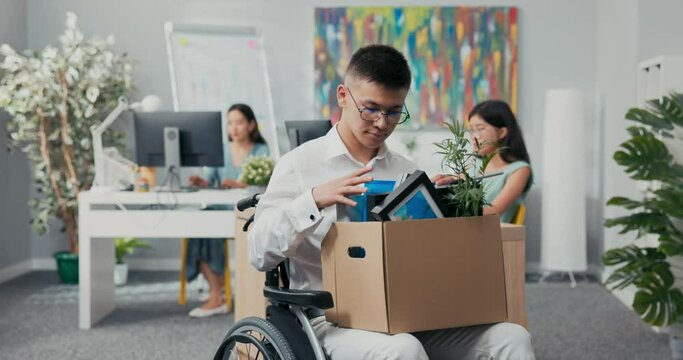 Young corporate office worker injured, accident, disabled, in wheelchair, holding a box of paraphernalia from his desk in lap, retiring, laid off, unemployed, changing positions