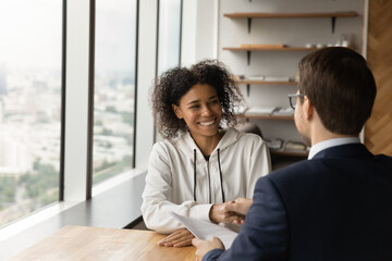 Happy friendly young female african american candidate shaking hands with male hr manager employer, accepting job offer, thanking for interview, making good first impression in modern office.