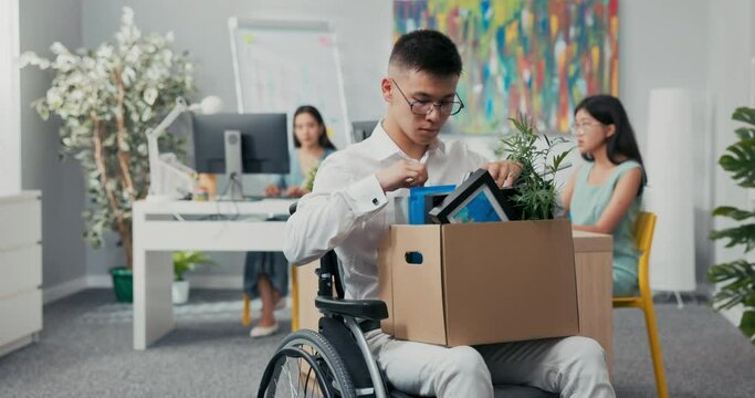 Young corporate office worker injured, accident, disabled, in wheelchair, holding a box of paraphernalia from his desk in lap, retiring, laid off, unemployed, changing positions