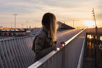 A girl in a trench coat stands on a modern bridge in the city and looks at the sunset. Tourist walks in Europe. Urban lifestyle