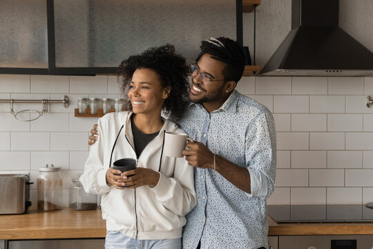 Happy Loving Affectionate Young African American Family Couple Standing In Modern Kitchen, Holding Cups Of Coffee In Hands, Enjoying Sweet Trusted Conversation, Daydreaming Visualizing Future.