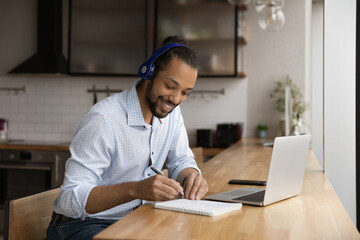 Happy young african american man in headphones writing notes in paper copybook, using computer...