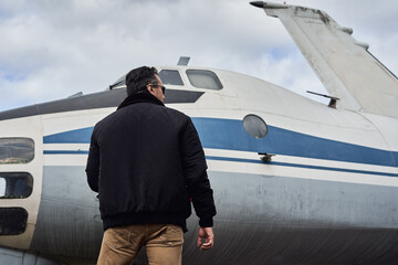 Fototapeta na wymiar Oldschool pilot in the bomber jacket posing near the aircraft, aviator male with scarf and sunglasses