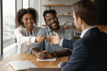 Happy young african mixed race family couple taking keys from male real estate agent. Smiling diverse husband wife feeling excited of purchasing own house or apartment, making deal with realtor.