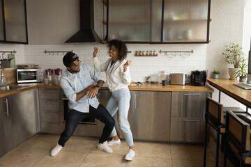 Sincere joyful laughing young african american family couple dancing in modern kitchen, listening energetic disco music, having fun entertaining spending carefree leisure time together at home.