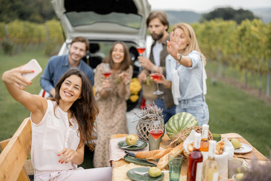Pleased european friends tasting local wine during picnic near vineyards in countryside. Woman making selfie photo on phone. Young men and women spending time together. Friendship, rest and leisure