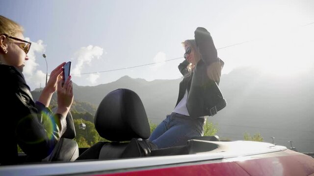 Beautiful girls take photos on a smartphone inside a red convertible. Posing and enjoying the rest among the mountains.