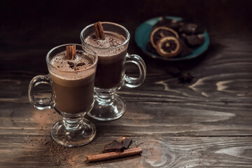 Two glasses of hot chocolate or coffee with cinnamon sticks on a wooden background. Warming coffee drink. Composition in dark style with copy space.