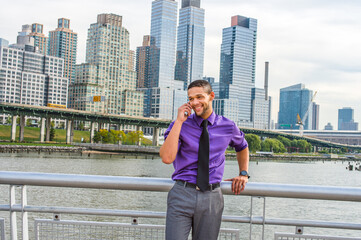 Fototapeta na wymiar Dressing in a purple shirt, gray pants, a black tie, a young businessman with a little beard and mustache is standing outside a busy business district, talking on his mobile phone.