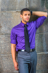 Obraz premium Dressing in a purple shirt, gray pants, a black tie, one hand putting in the back of his head, a young college student with a little beard and mustache is leaning against the wall, thinking outside..