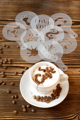 Top view of ceramic cup of hot cappuccino coffee latte with drawing picture of cafe sign with cinnamon or cocoa on milk foam, different stencils, coffee beans on wooden table