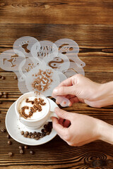 Top view of ceramic cup of hot cappuccino coffee latte with drawing picture of cafe sign with cinnamon or cocoa on milk foam, woman hands holding different stencils, coffee beans on wooden table