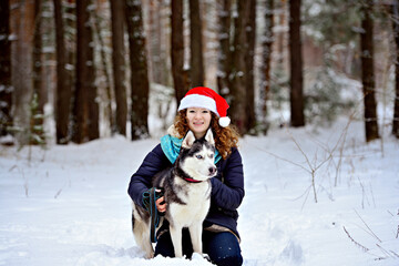 Fototapeta na wymiar Young laughing girl in a Christmas hat with a Husky dog in the winter forest. She sits and hugs her dog. She looks into the camera lens. Horizontal orientation.