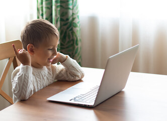 Little boy in a white sweater is sitting at a laptop and speaking by video call. 