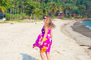 An adult blonde in a bright dress that sways in the wind, on the tropical seashore. Travel and tourism.