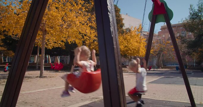 Children Having Fun, Swinging On Seesaw Board, Playing Playground In Park. Concept Of Happy Childhood, Positive Emotions. Baby Boy And Girl Riding On Swing Outdoors. Mother Pushing Child On A Swing