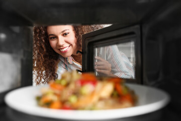 Young African-American woman heating food in microwave oven, view from inside - obrazy, fototapety, plakaty