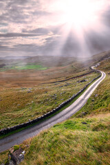 Small narrow country road on a hill by a traditional stone wall with beautiful nature scene. West of Ireland. Irish landscape. Green grass. Fog in the background. Transportation industry. Sun flare