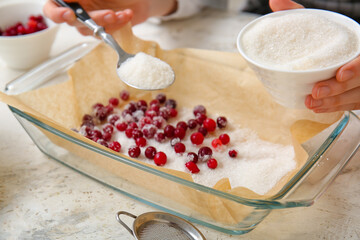 Woman sprinkling sugar onto cranberry on light background, closeup