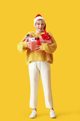 Surprised young woman in Santa hat and with Christmas gifts on color background