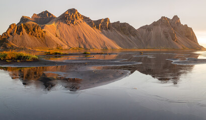 Color landscape photo of Vestrahorn mountain and reflection in Iceland. Photo is at sunrise. Mountain is bathed in golden light and reflected in tidal plain.