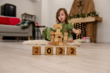 Wooden cubes with the numbers 2022 stand on the floor in a row. Background of a blurred happy girl in a green dress, who is playing, building a tower from a construction set. Child development at home