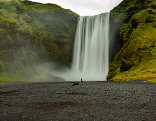 Closeup of Skogafoss waterfall in Iceland at sunrise.  Black and gray gravel in foreground. Green cliffs, one single person at base of waterfall.