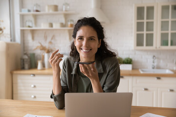 Smiling Latina woman sit at table with laptop holds smartphone talks on speakerphone, leave voicemail message chatting remotely enjoy personal or formal distant conversation. Modern ai tech concept - obrazy, fototapety, plakaty
